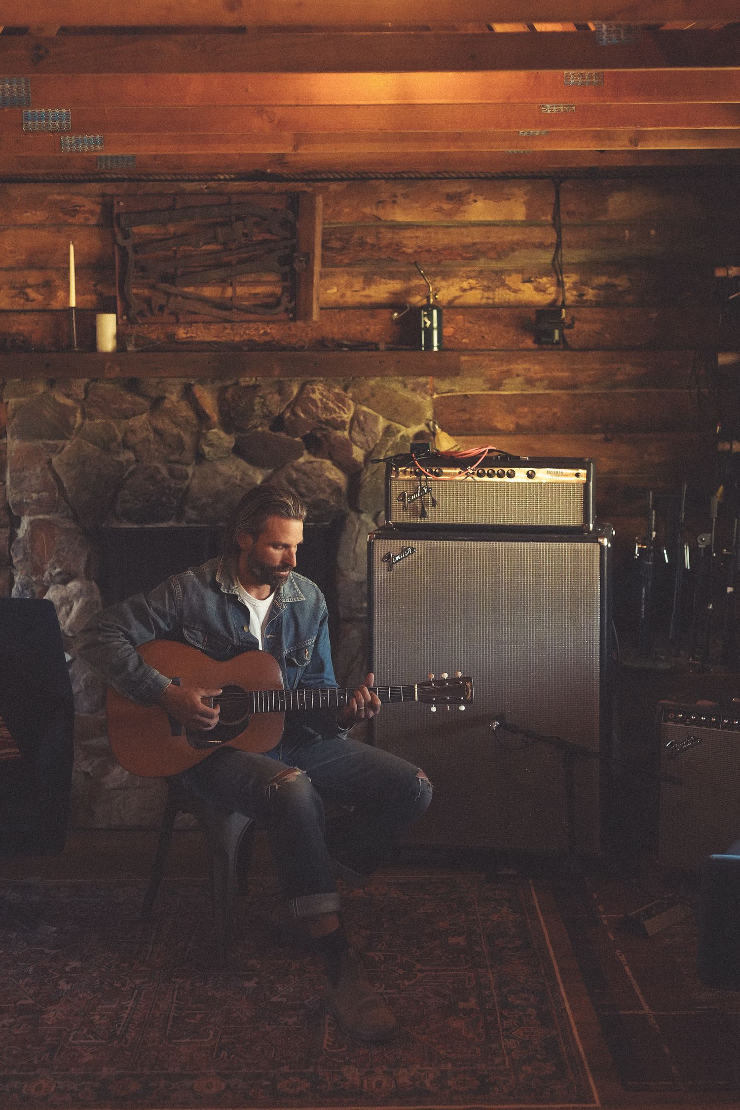 A musician plays acoustic guitar while sitting on a wooden bench in a dimly lit rustic room with stone walls.