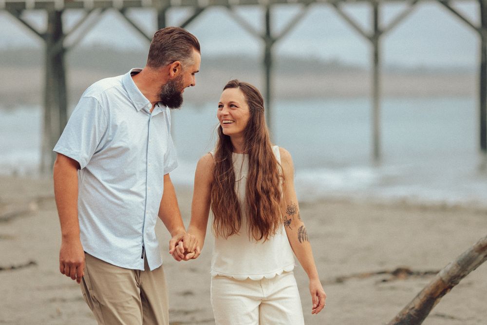 Couple holding hands and smiling at each other while walking on a beach near a pier.