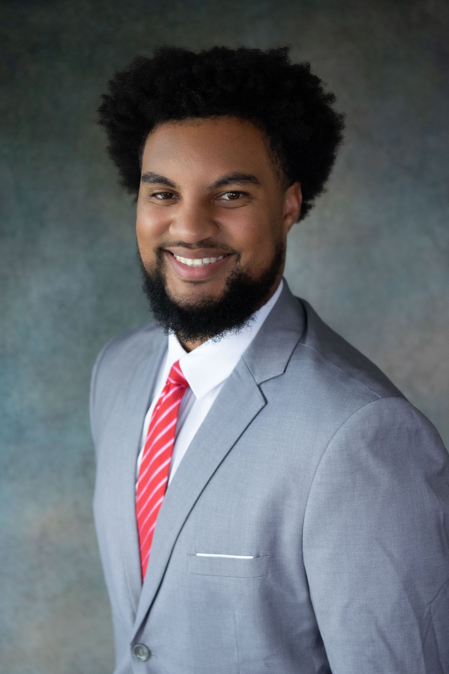 Professional headshot of person in grey suit and red tie against studio background.