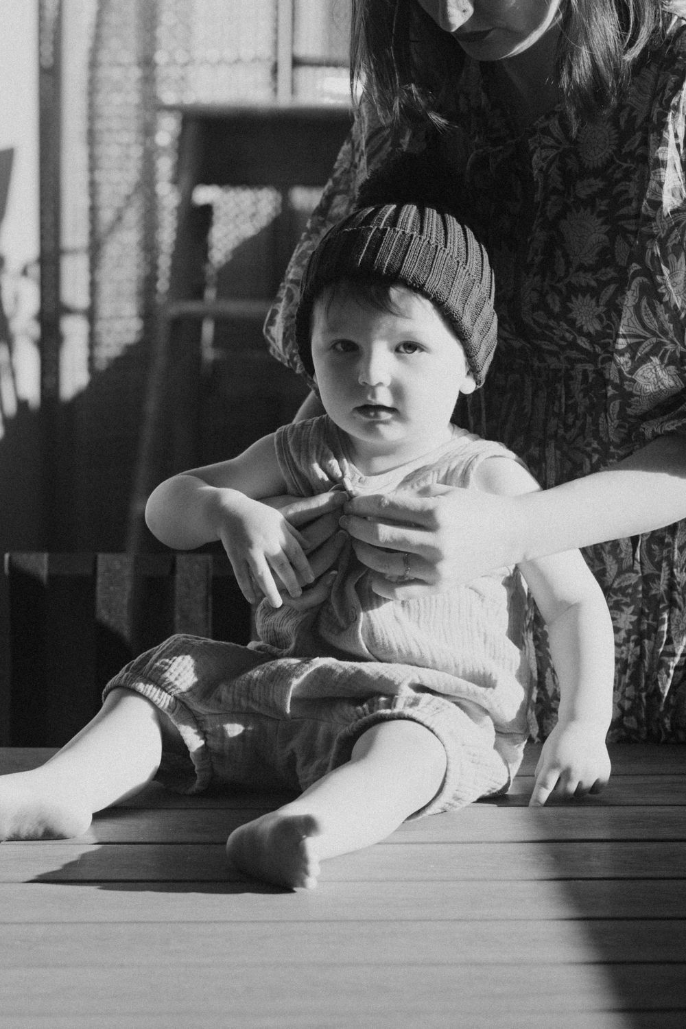 A young child in a knit hat sits on a wooden deck in a casual black and white portrait.