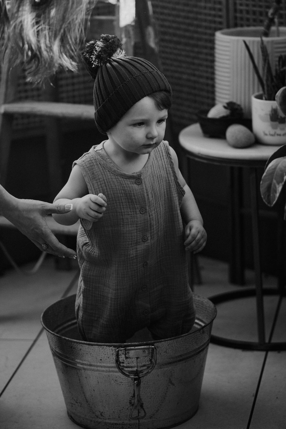 Child in knit hat and romper stands in vintage metal tub during indoor photoshoot.