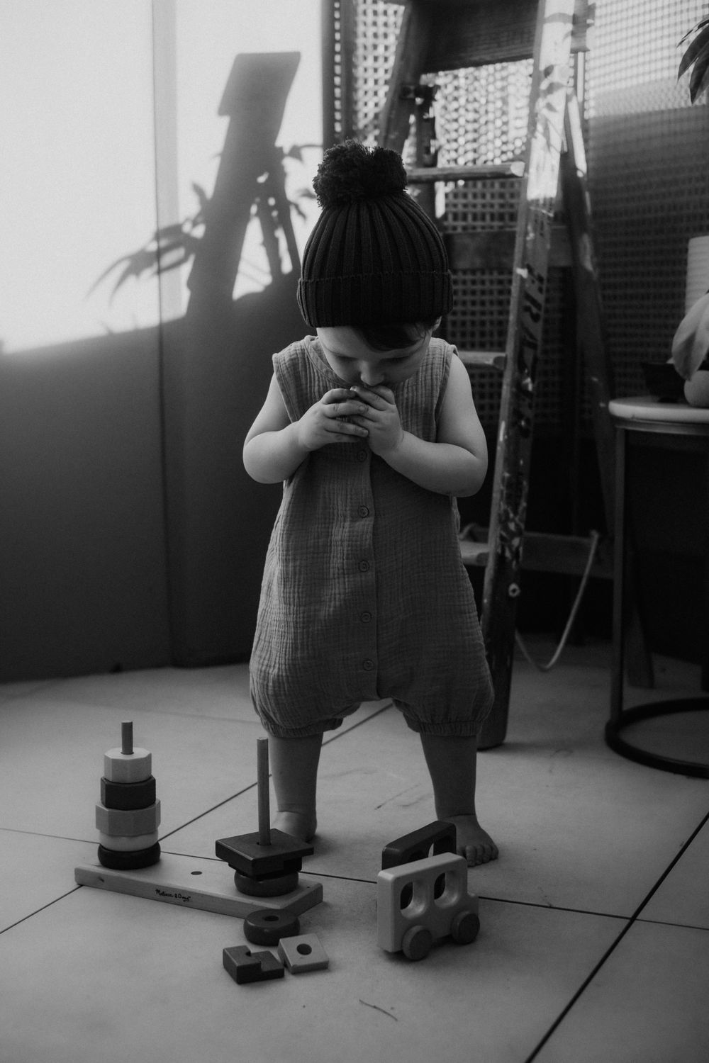 Child in gray romper and knit beanie plays with toys on tiled floor.