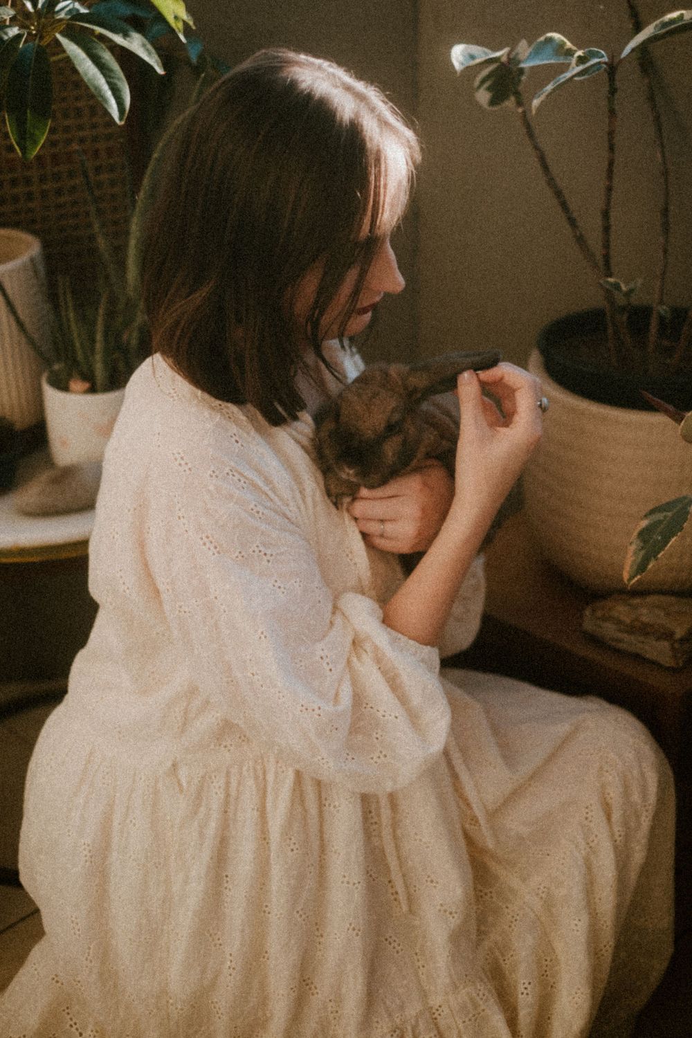 A child in a white flowing dress sits indoors near potted plants.