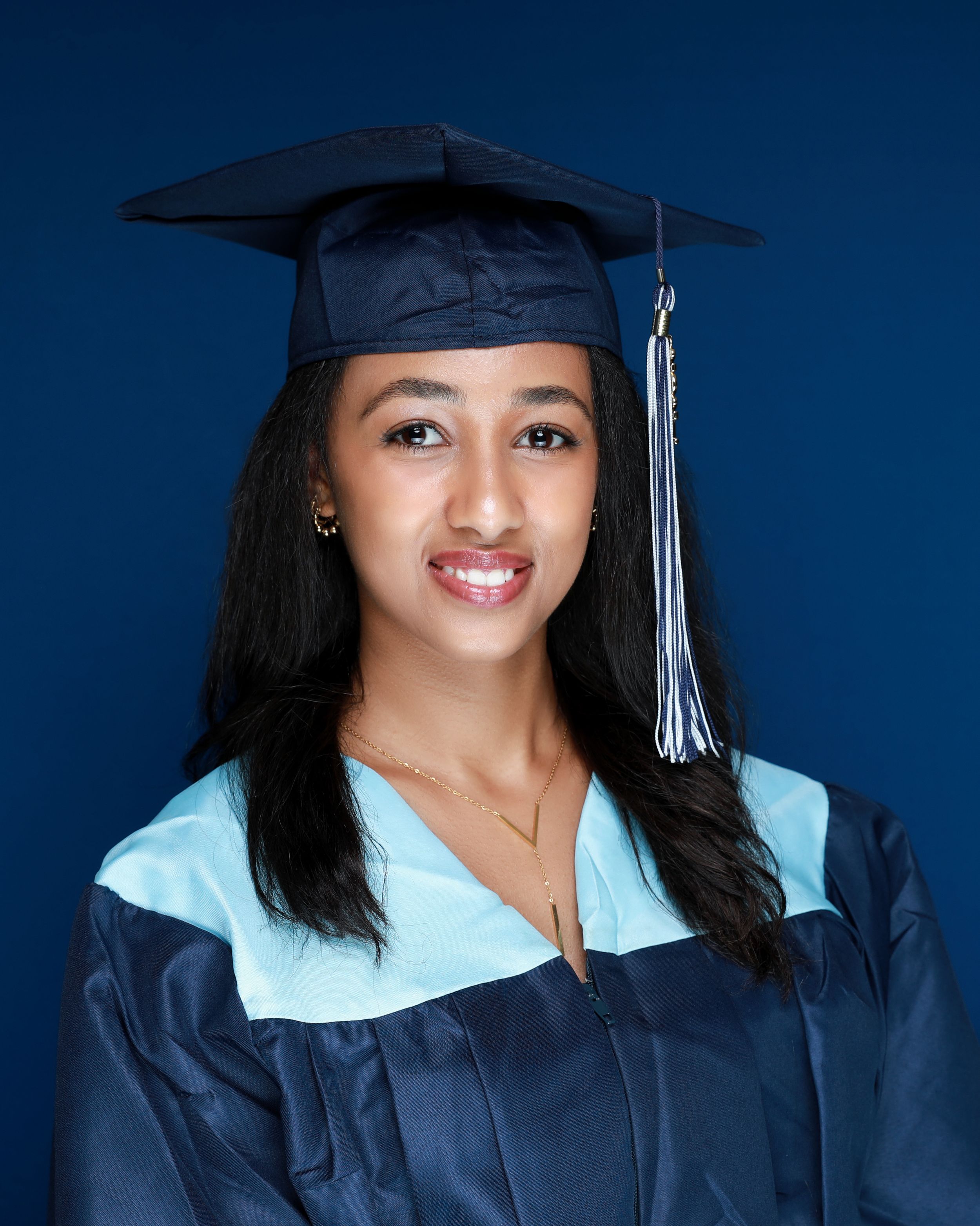 Graduate wearing navy blue graduation gown and cap with silver tassel against dark blue backdrop.