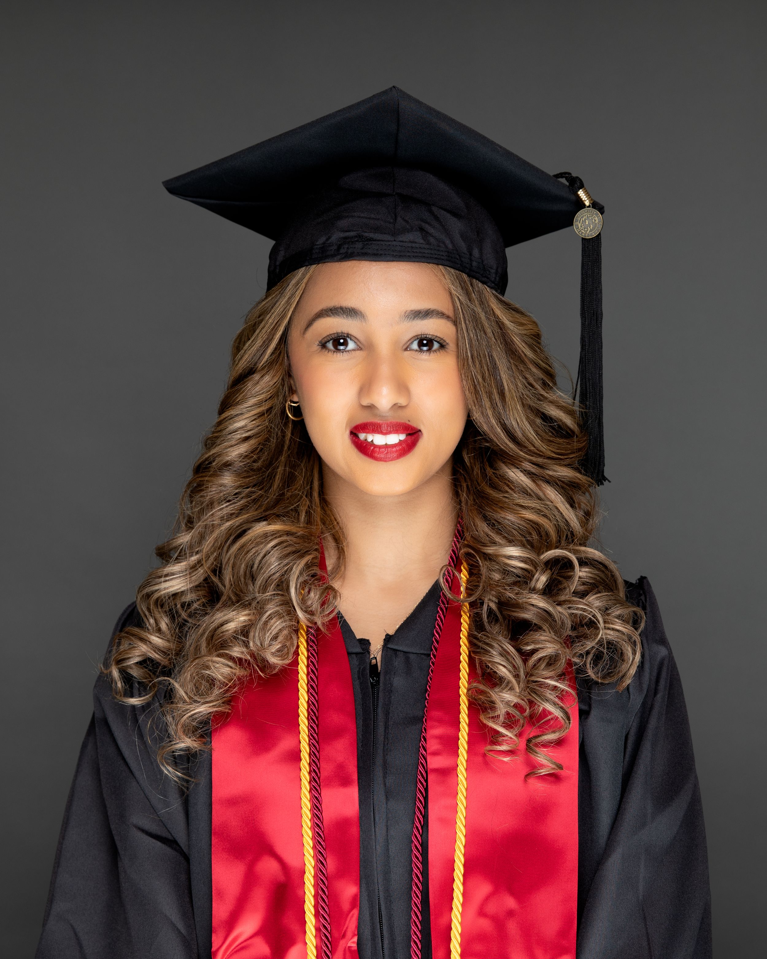 Young woman in black graduation cap and gown with red sash smiling for her graduation portrait photo.