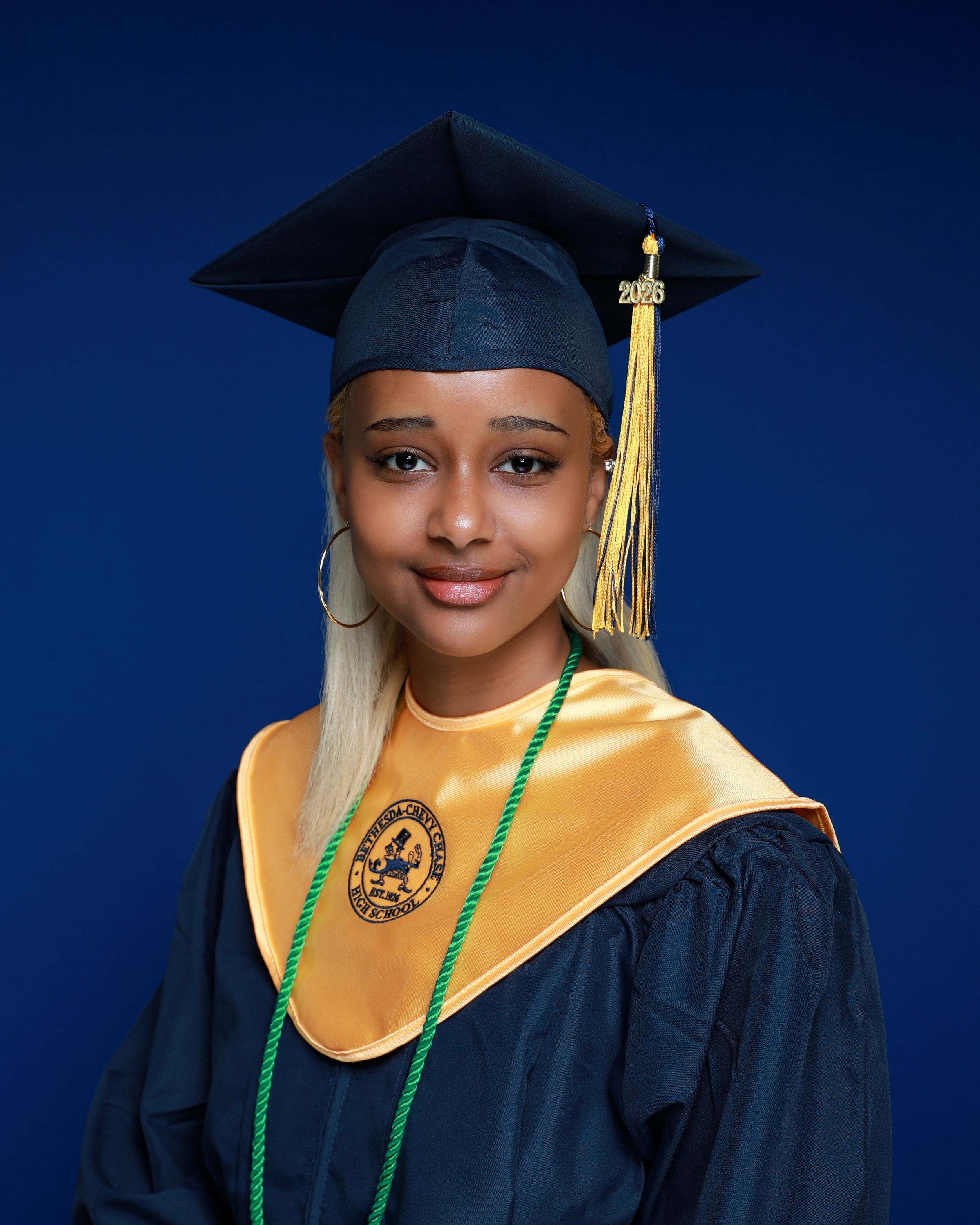 Young woman in navy graduation cap and gown with gold hood and green honor cord poses for portrait.