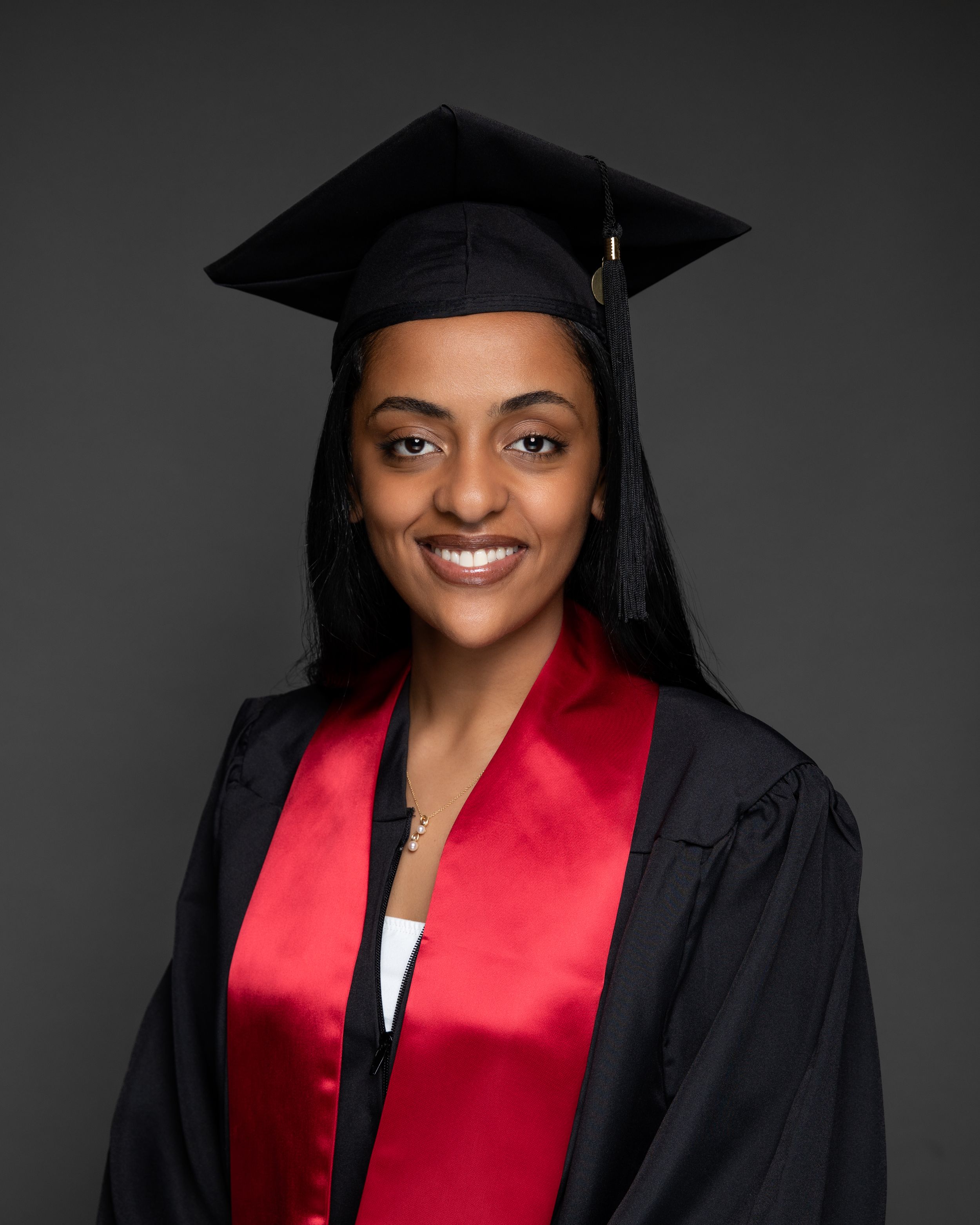 Young woman smiling in black graduation cap and gown with red sash, posing for professional graduation portrait.