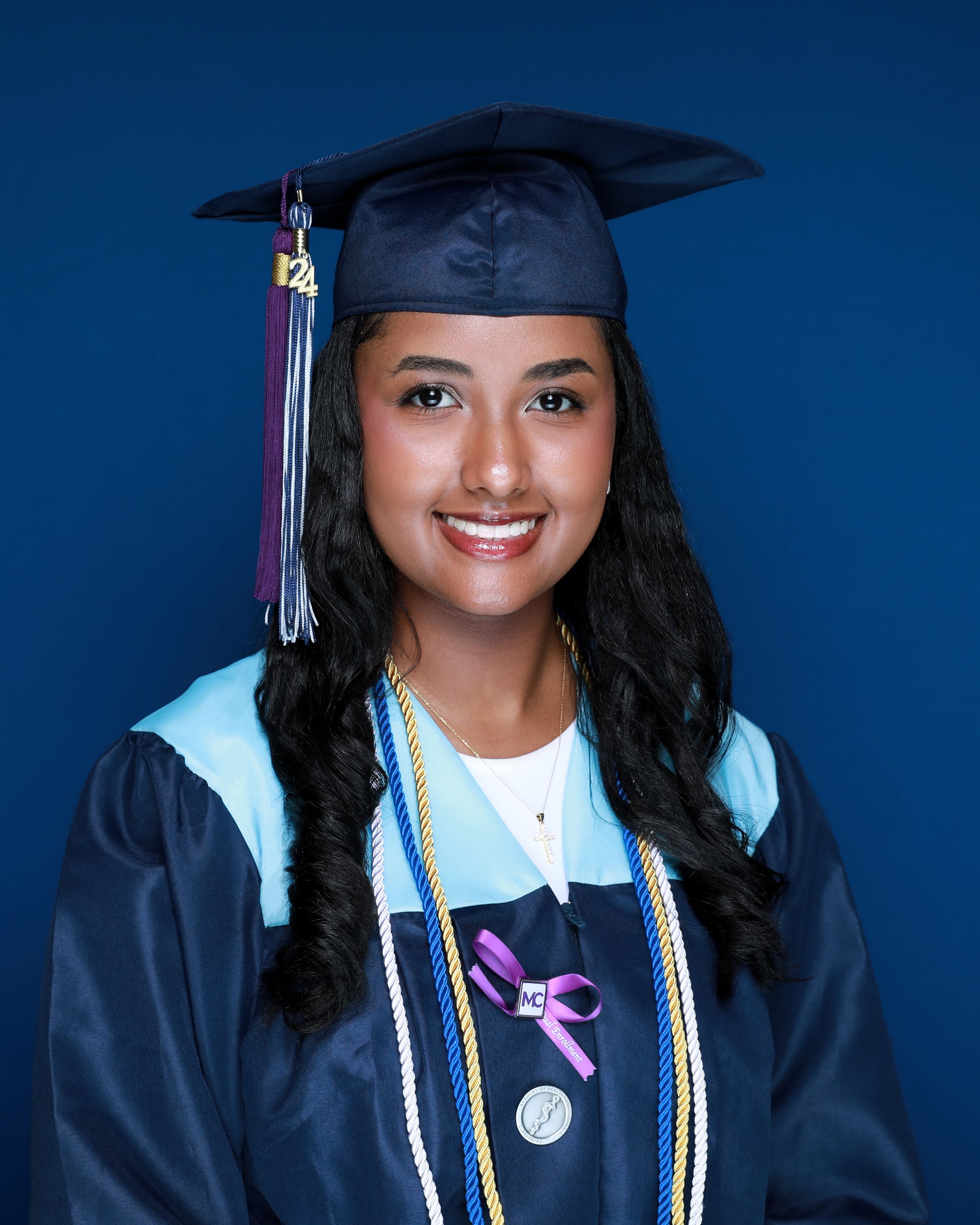 Graduate in navy and light blue cap and gown with tassel and honor cords against blue background.