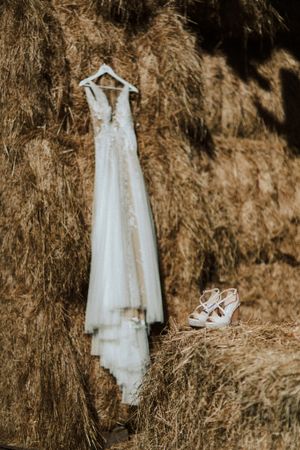 Wedding dress and shoes hanging on rustic hay bale background.
