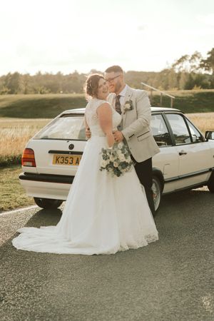 A classic white vintage car serves as backdrop for elegant wedding photos in a rustic outdoor setting.