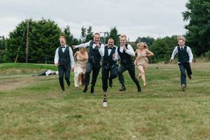 Wedding party runs and jumps playfully across a green grass field on a summer day.