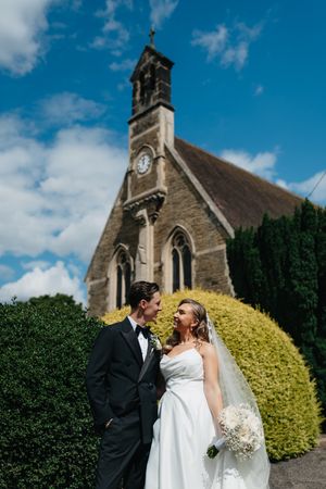A newlywed couple shares a moment outside a charming stone church with Gothic architecture on a sunny day.