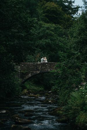 A historic stone bridge crosses a rushing stream in a lush forest setting with figures sitting together on the arch.