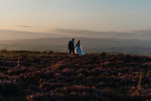Two figures stand on a hilltop silhouetted against a sunset sky, surrounded by purple heather blooming on moorland.