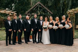 A wedding party in formal black attire and white gowns poses in front of trees during an outdoor photo session.