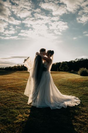 A silhouetted couple in wedding attire embrace on a golden hilltop at sunset, their veil flowing dramatically in the breeze.