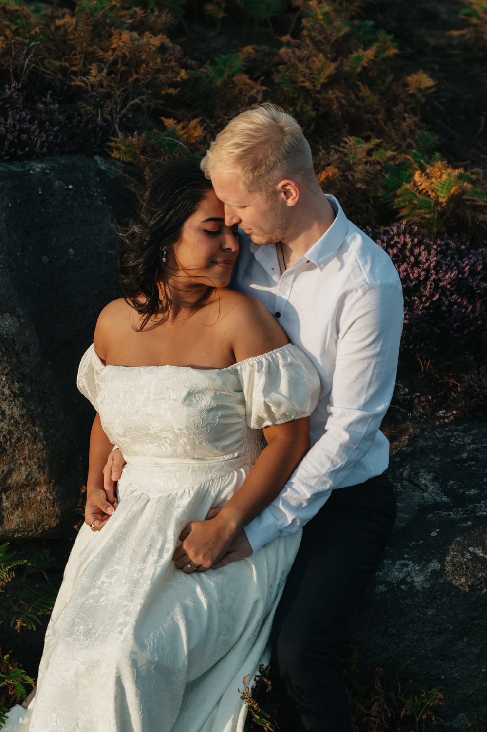 Romantic couple embraces in an outdoor wooded setting wearing white wedding attire at sunset.