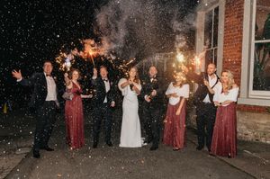 Wedding party celebrates outside at night with sparklers creating a magical atmosphere against brick building backdrop.