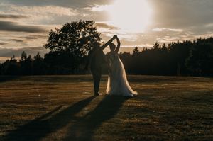 Wedding couple stands in silhouette against a dramatic sunset sky on a grassy hilltop, creating a romantic atmosphere.