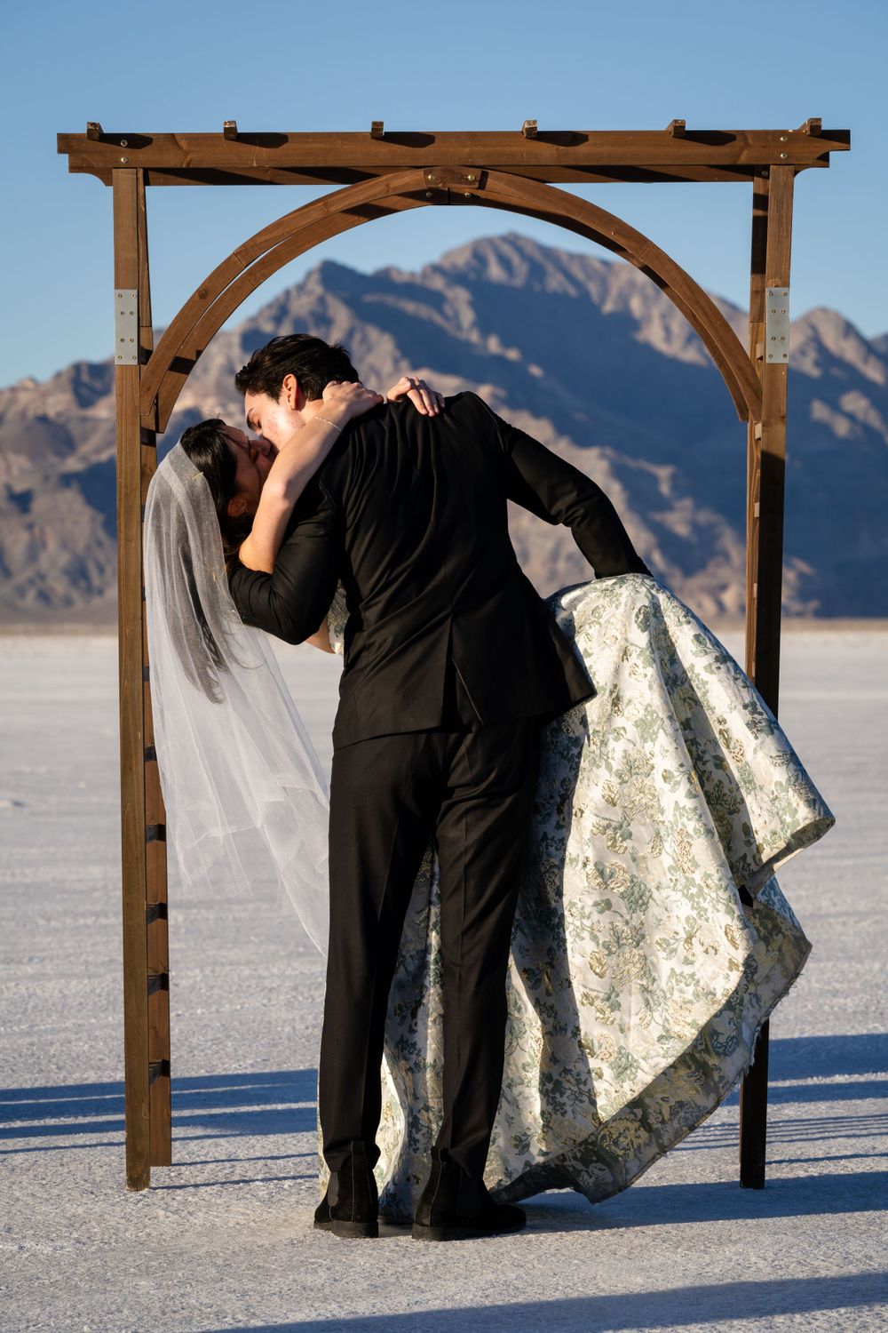 Wedding couple embracing under wooden arch on salt flats with mountain backdrop.