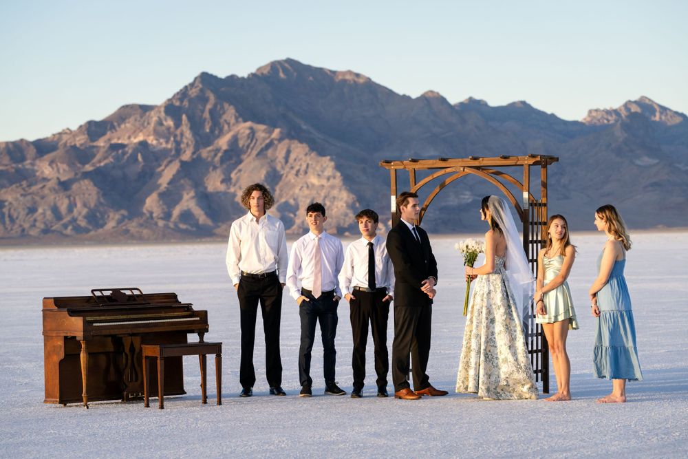 Wedding party posed with piano on salt flats with mountain range in background.