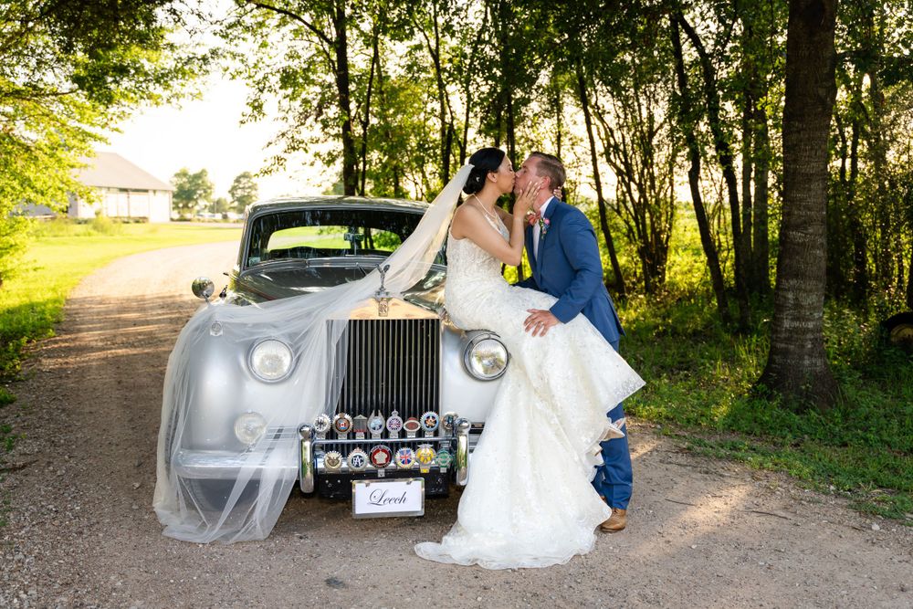 Romantic wedding couple poses with classic white Rolls Royce on tree-lined path.