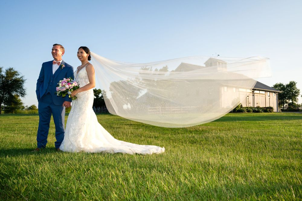 Wedding couple stands in green field as bride's veil dramatically flows in the wind.