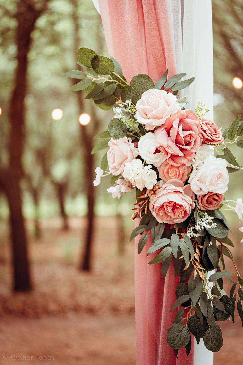 Pink roses and greenery decorate a coral-colored fabric drape in a wooded outdoor setting with twinkling lights.