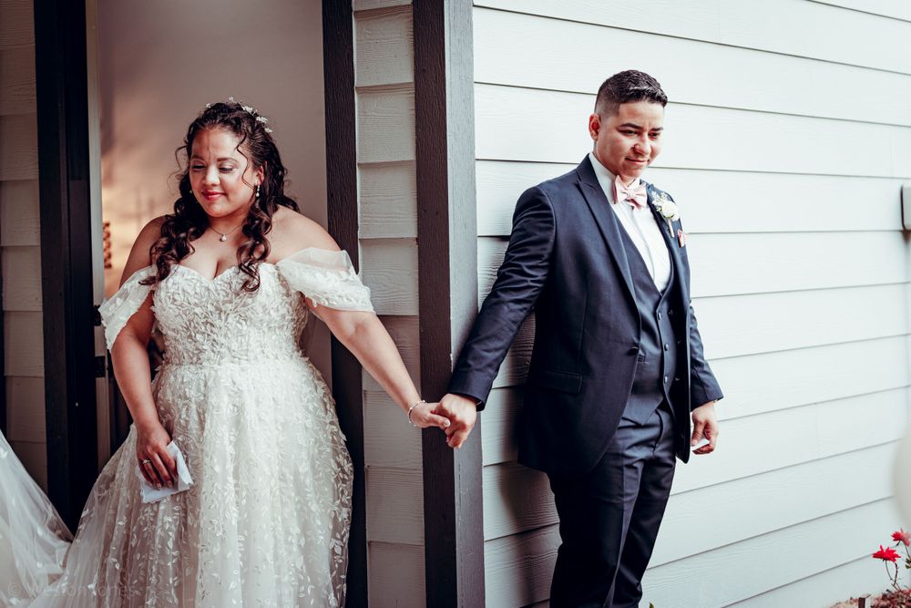 A couple in wedding attire holds hands while standing against a white wall.