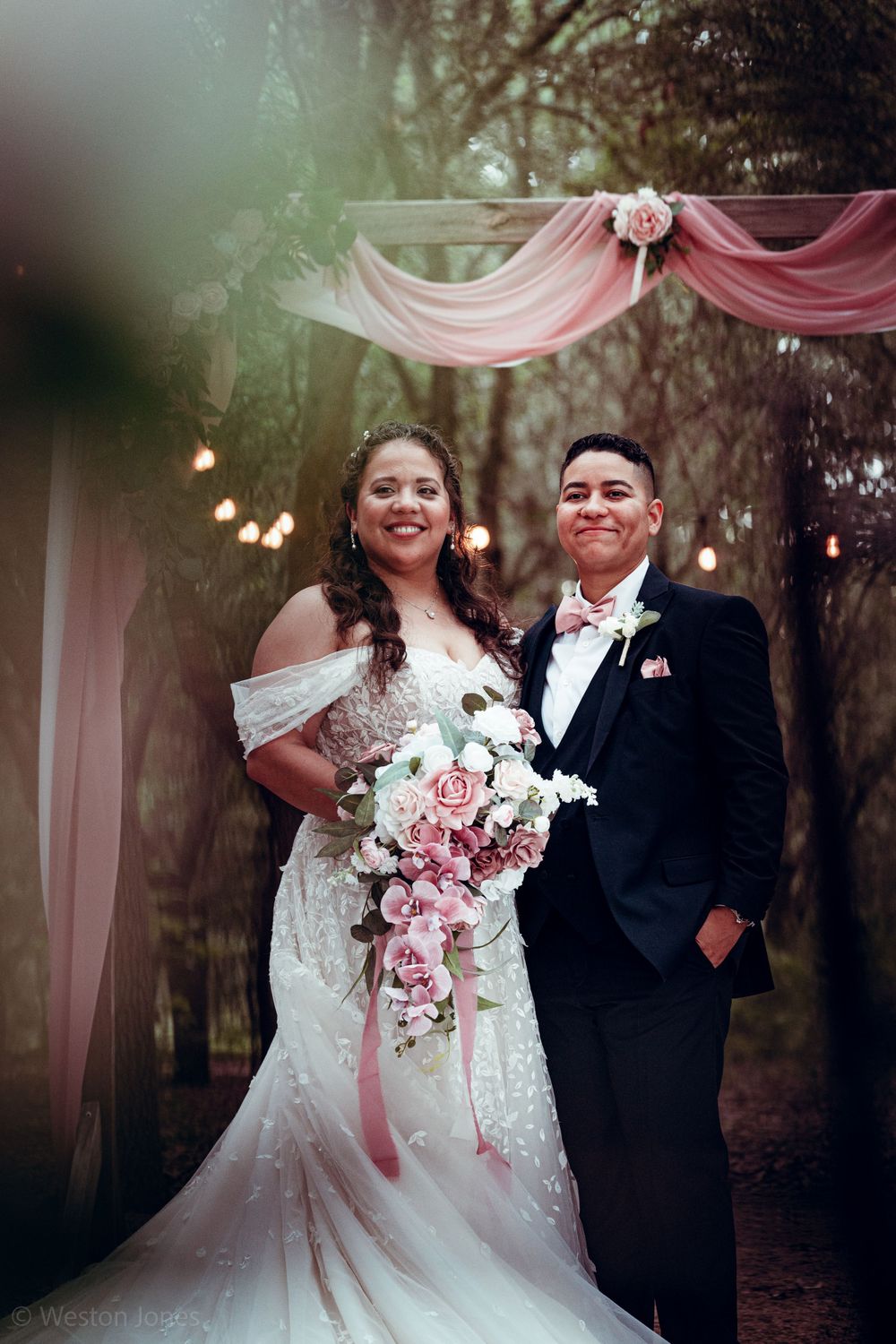 A couple poses under a pink draped arch decorated with roses in a romantic forest setting.