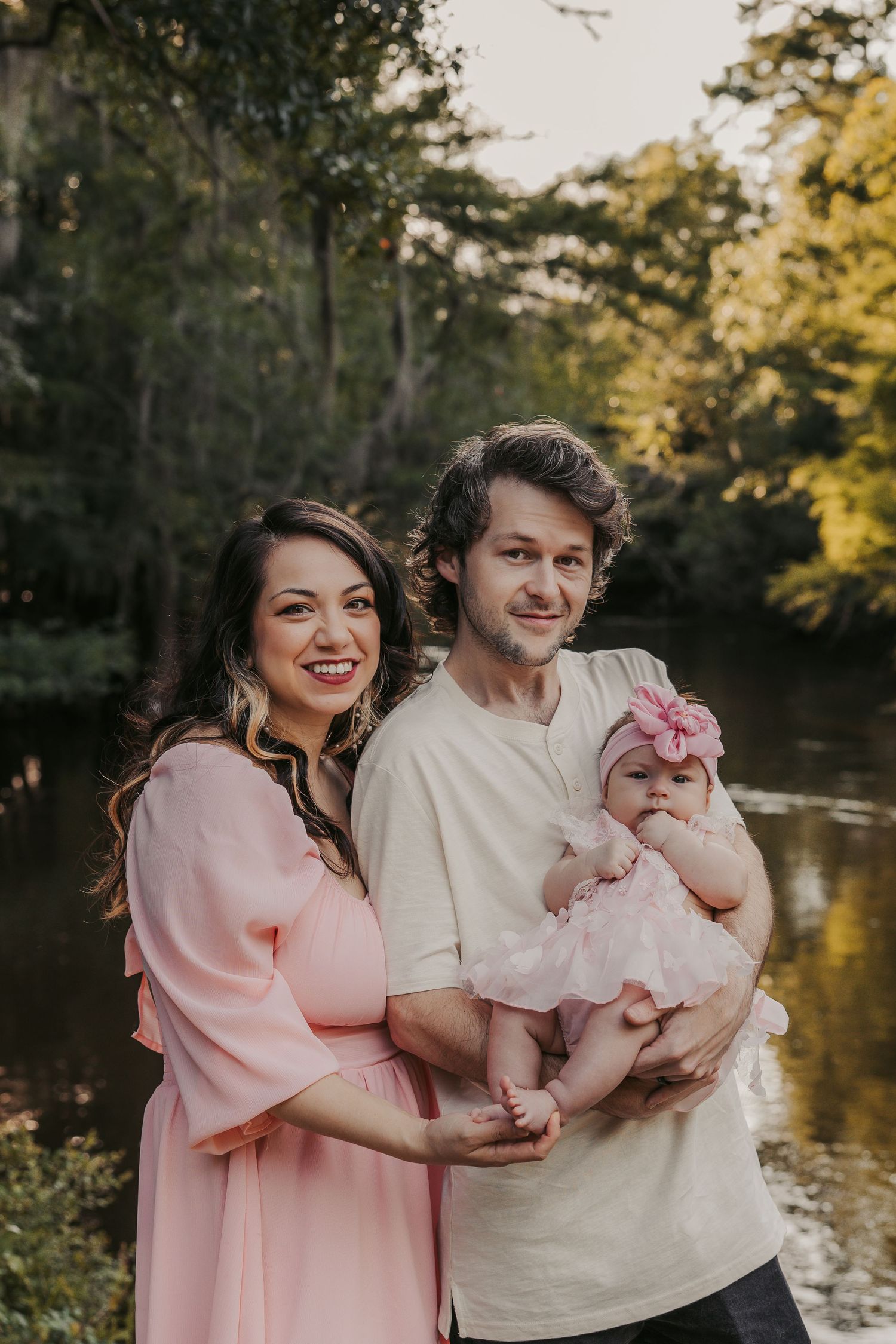 Family portrait series by a lake with parents in pink and white outfits holding their infant child.