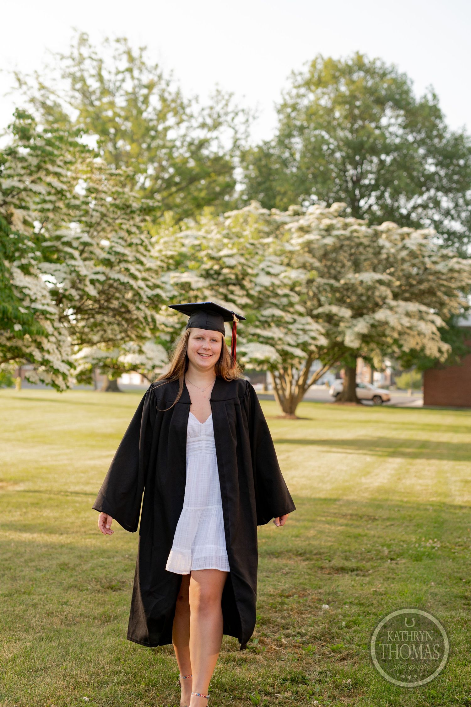 Graduate poses in traditional black graduation attire beneath flowering trees on a grassy lawn.