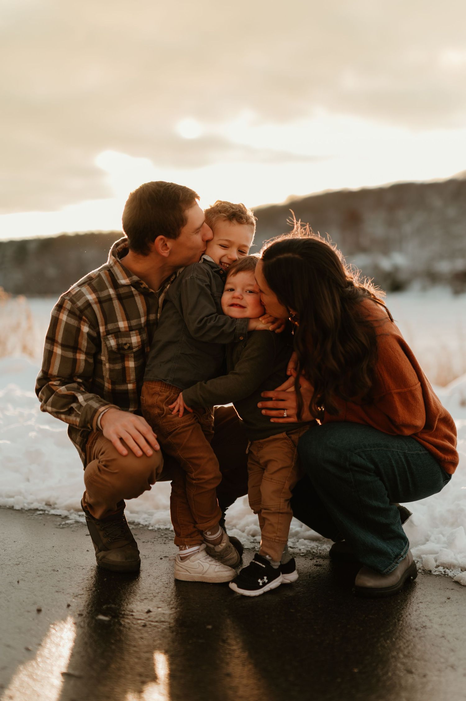 Family sharing kisses outdoors in snowy winter setting at golden hour, parents embracing two young children during session in West Bend, WI.