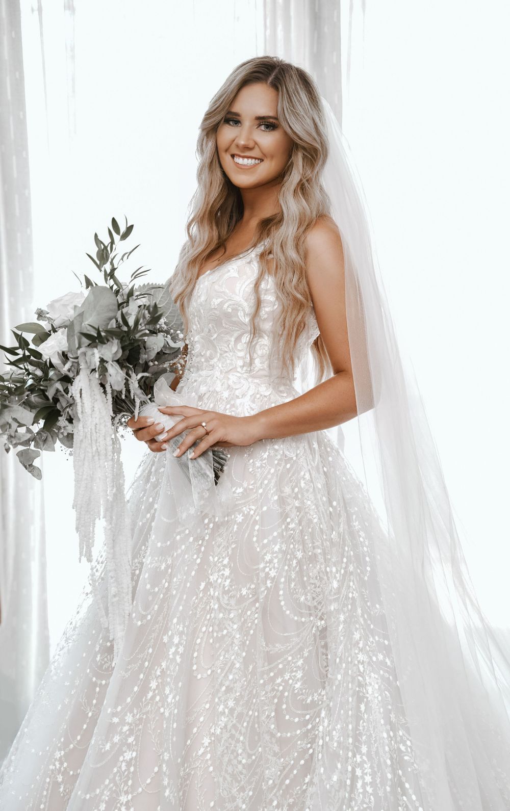 Bride in sparkly lace wedding gown holds bouquet with eucalyptus and white veil.