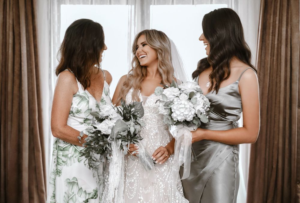 Three women in formal attire hold white floral bouquets while sharing laughs.
