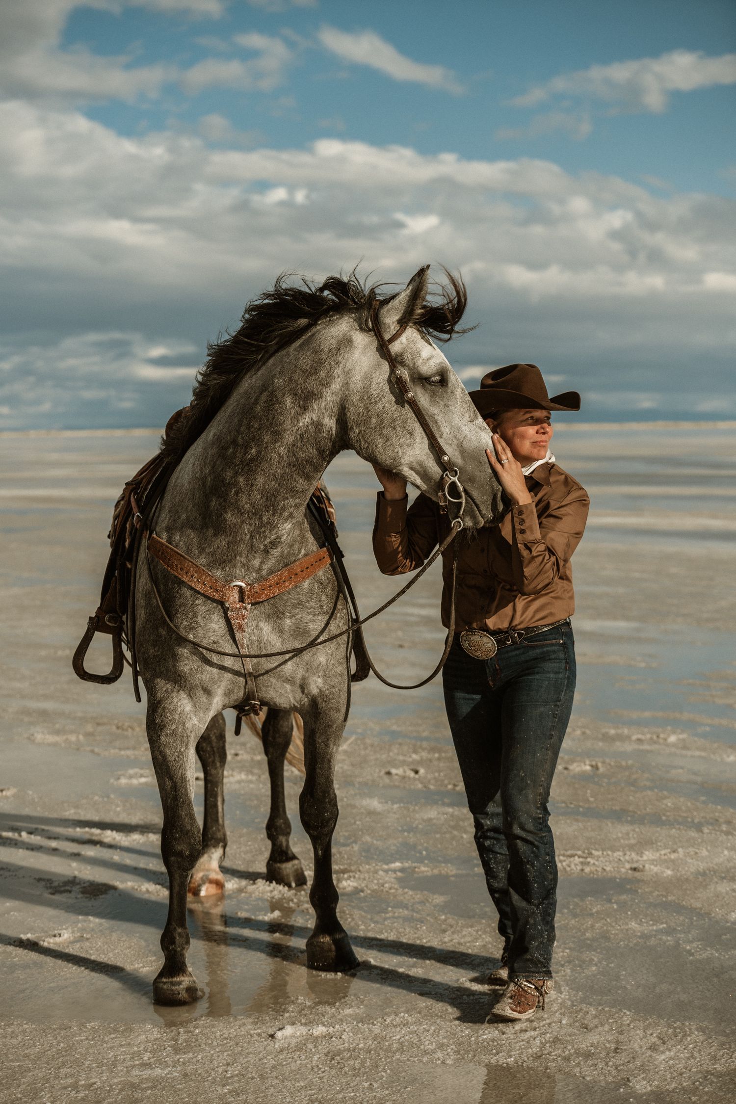 Person in western wear stands with gray horse against desert landscape and cloudy sky.