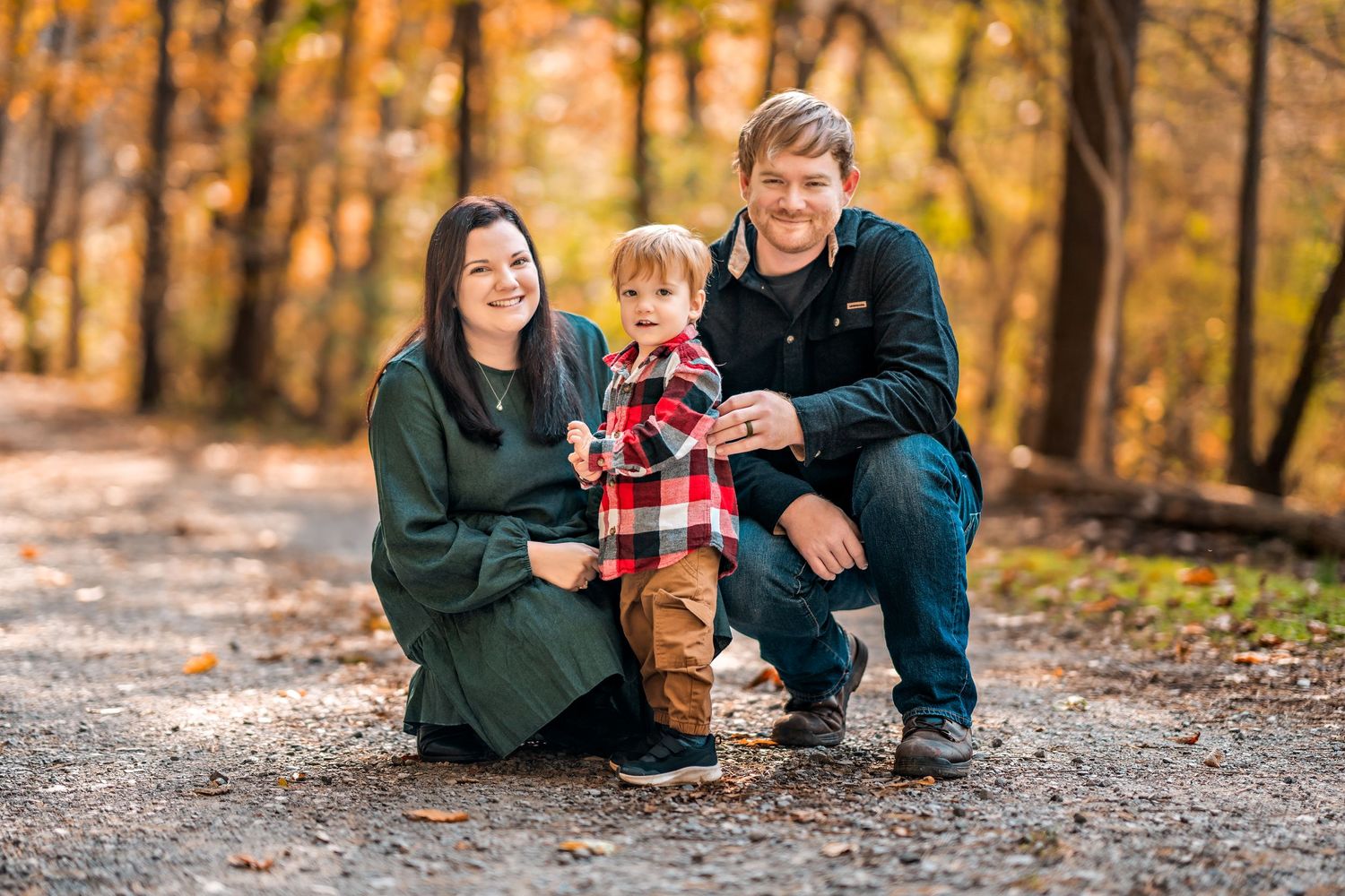 A family in matching green and plaid outfits poses together on a hiking trail surrounded by autumn foliage.