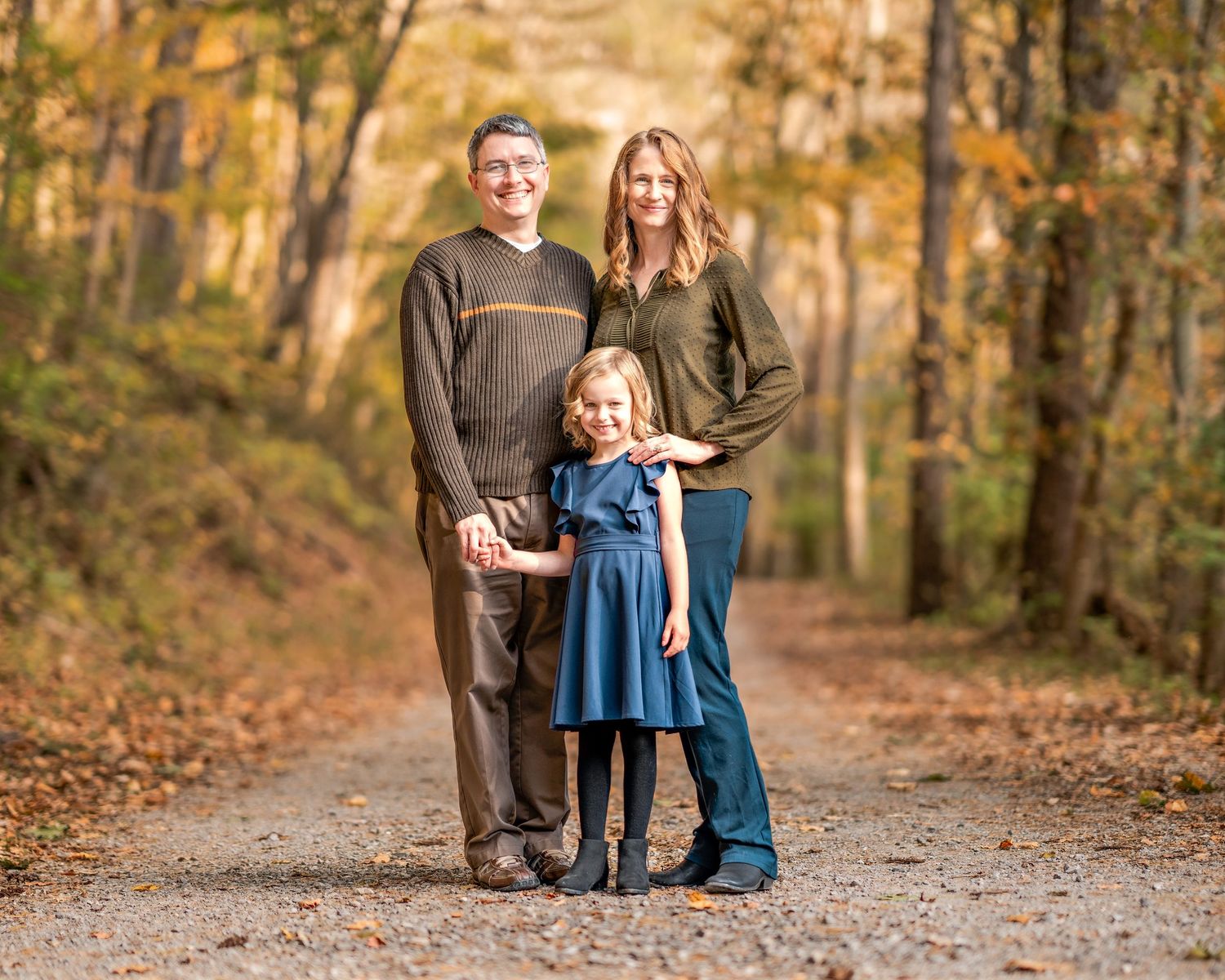 Family of three poses on an autumn trail surrounded by golden trees and fallen leaves on a dirt path.