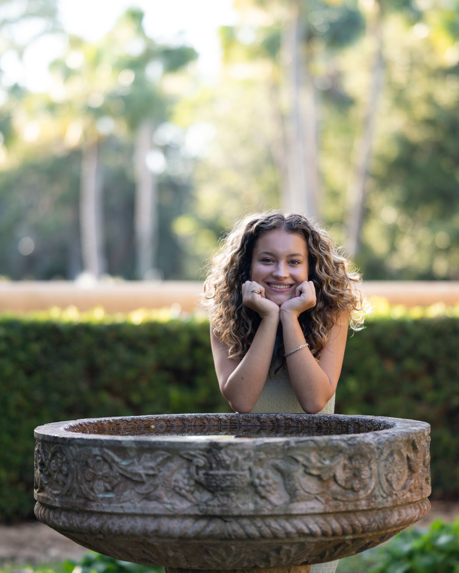 A curly-haired figure rests their chin on hands while posing playfully in a large ornate stone garden fountain basin.
