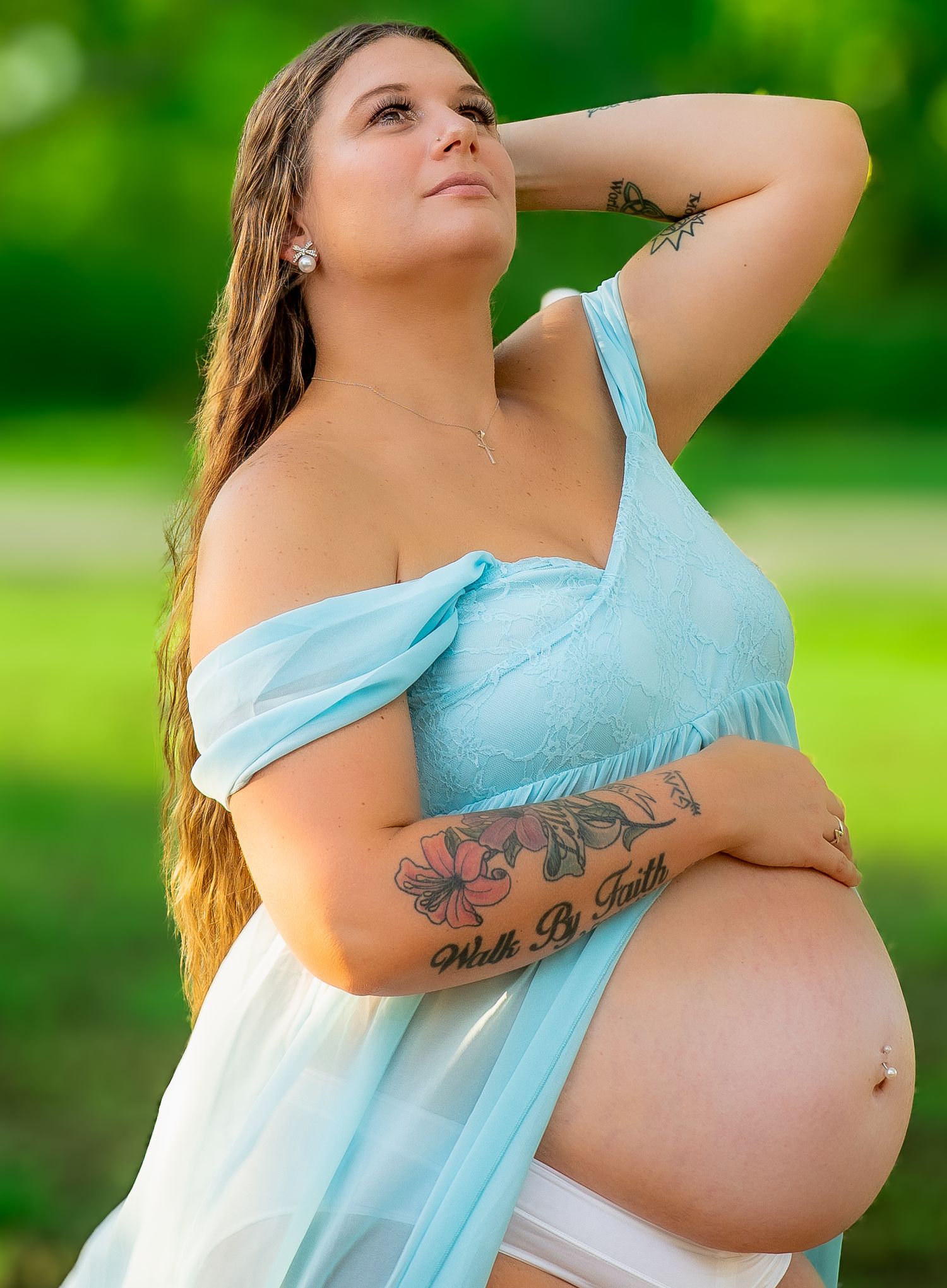 A pregnant person in a light blue flowy dress poses outdoors against a blurred green background.