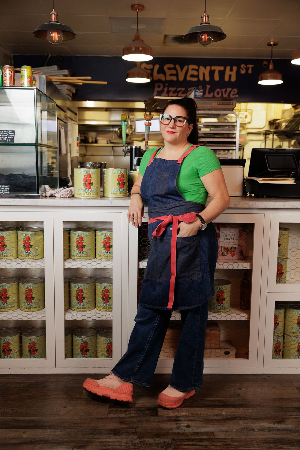 Chef Nicole Votano in denim apron and green shirt stands in front of Seventh St Pizza counter with vintage-style lighting.