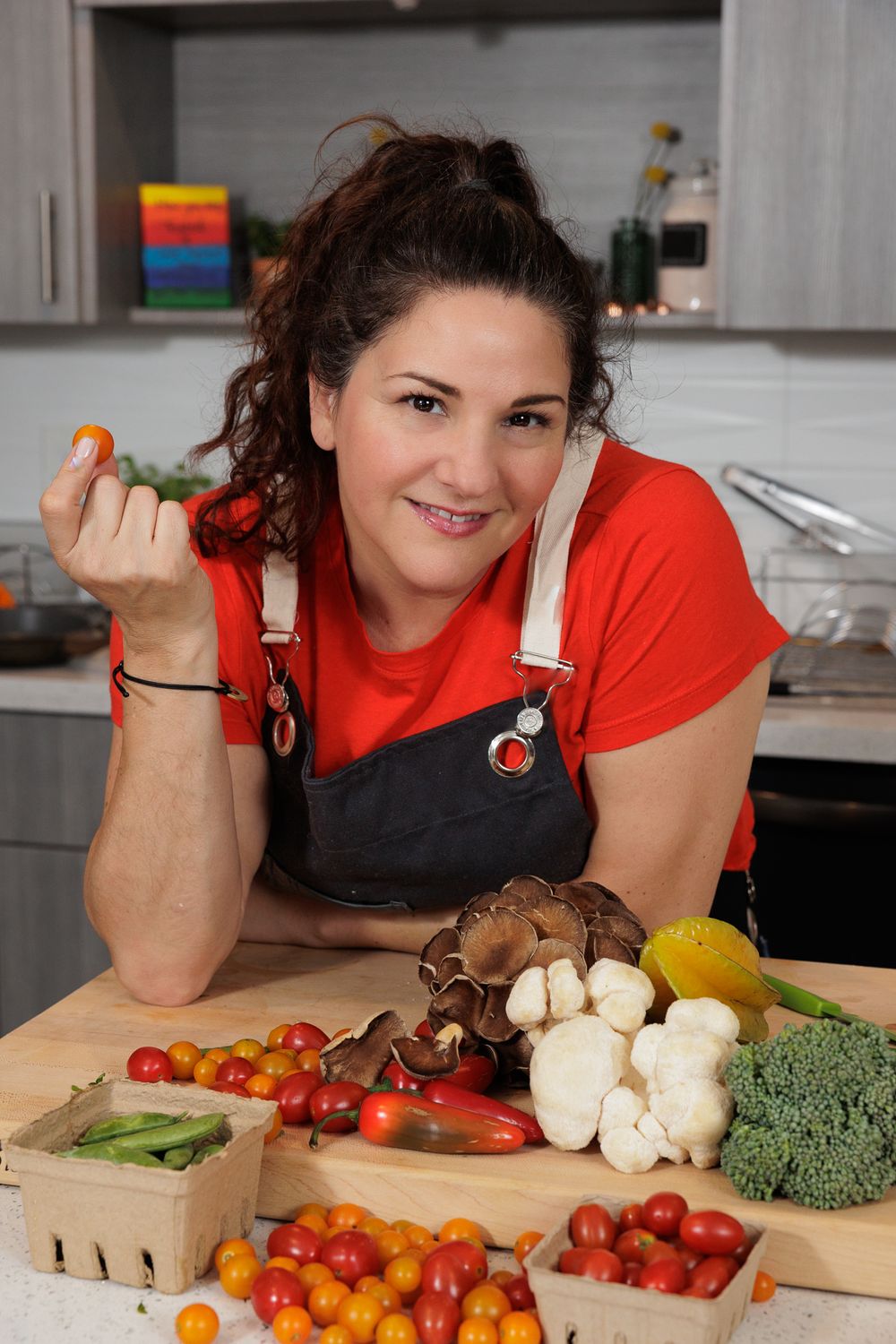 Chef Nicole Votano in red shirt and overalls poses at kitchen counter with fresh vegetables and mushrooms arranged on wooden crates.