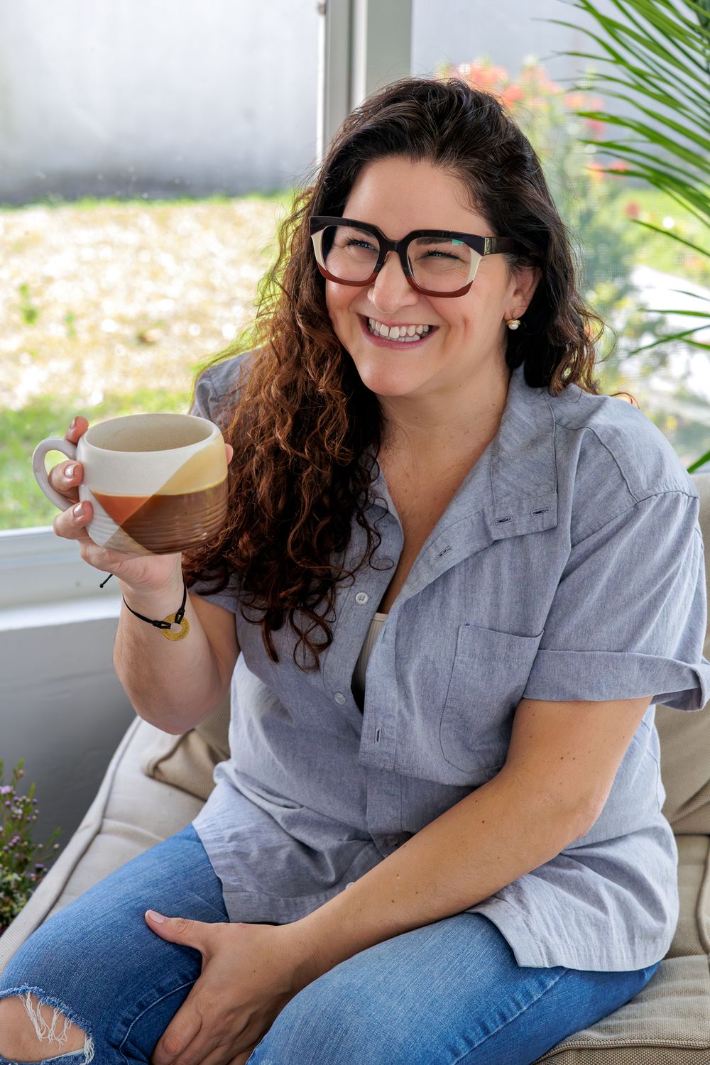 Chef Nicole Votano wearing glasses and a gray button-up shirt sits with a coffee cup near a window with plants.