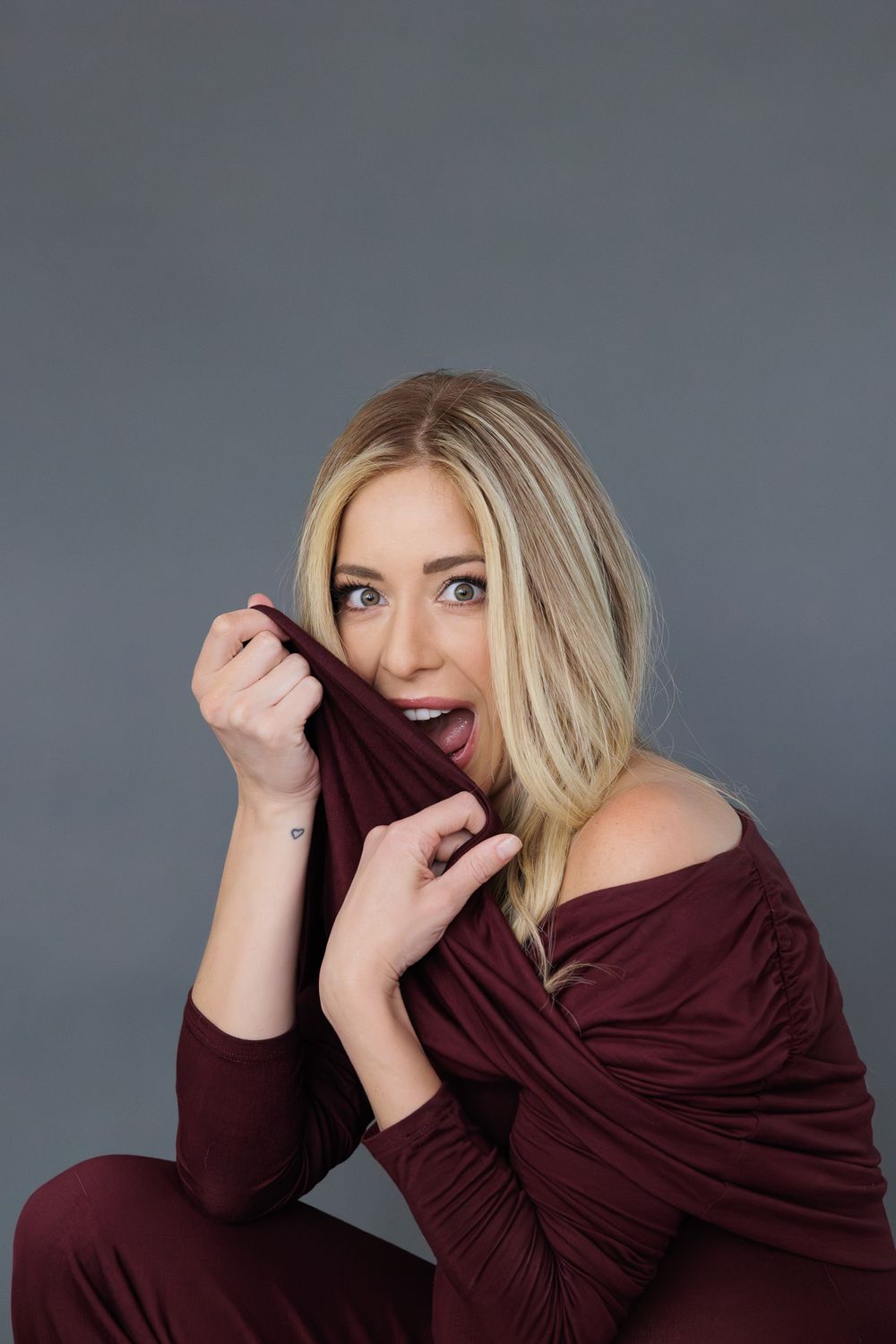 A woman, Carmen Aub, is in a burgundy off-shoulder outfit poses playfully against a gray backdrop.