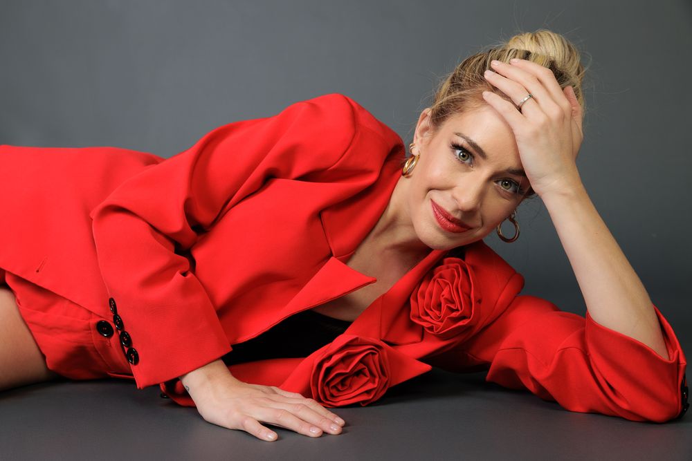 A woman, Carmen Aub, in a vibrant red suit lies on their side against a dark gray backdrop in a fashion pose.