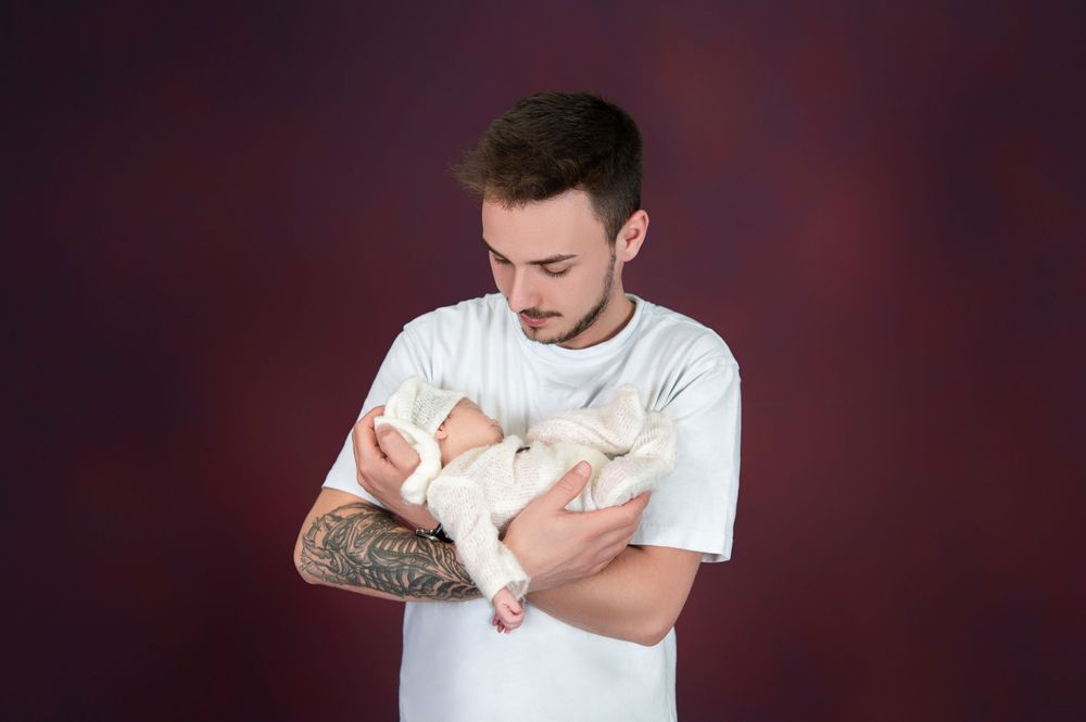 Parent holding sleeping newborn baby against dark burgundy backdrop.