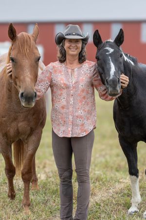 Horse handler stands between a chestnut and black and white horse against a red barn backdrop.