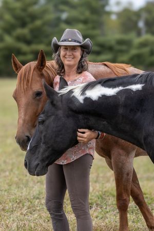 A person in cowboy hat stands with two horses - one brown and one black with white markings in a grassy field.