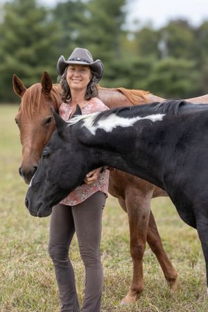 A happy rider stands with two horses in a field, one dark and one brown, wearing western attire and a cowboy hat.