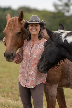 Two horses flank someone wearing a cowboy hat and floral shirt in a rural outdoor setting.